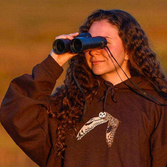 Person wearing a brown hoodie with a logo, looking through binoculars in a field.