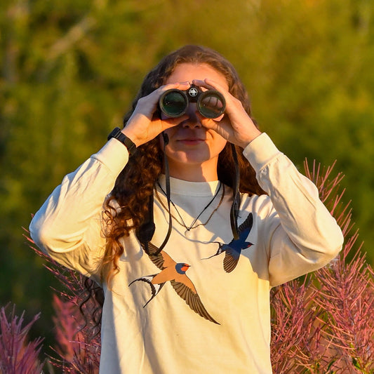 Person using binoculars in a natural setting with greenery and pink plants.